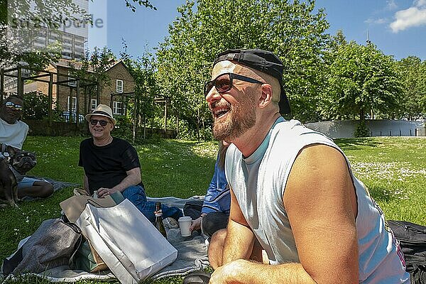 Man in sunglasses and headband laughing during a sunny park picnic with friends and a dog in the background