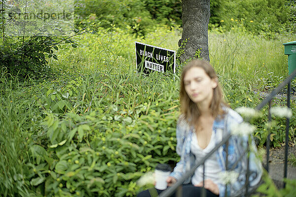 A young adult in the foreground is out of focus  with a Black Lives Matter sign in clear view amongst lush greenery in the background.