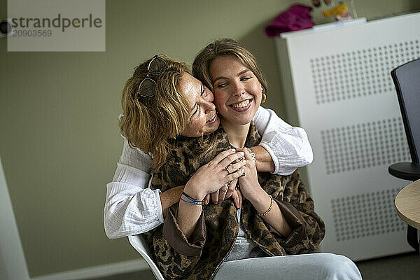 Two women share a joyful hug with one resting her head on the other's shoulder  both smiling warmly in an indoor setting.