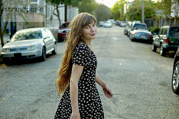 Woman with long hair wearing a polka dot dress smiles over her shoulder while walking down a residential street lined with parked cars.