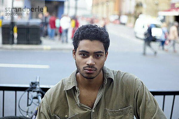 A young man with curly hair looks calmly at the camera  with a blurred city street scene in the background.