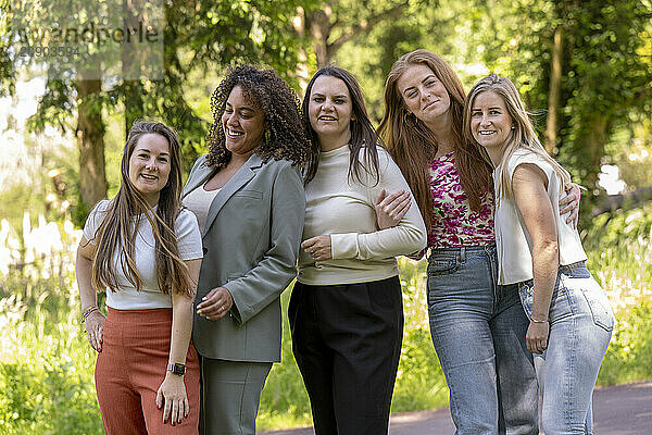 Group of five smiling women posing together in a sunny park setting.