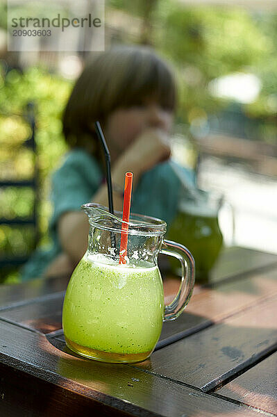 Refreshing limeade in a pitcher with a red straw on a wooden table  with a blurred child in the background.