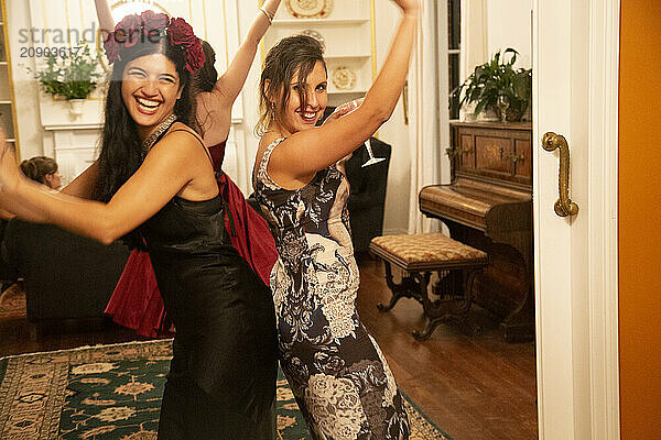 Two women dressed in elegant evening gowns dancing and laughing together in a vintage-style room.