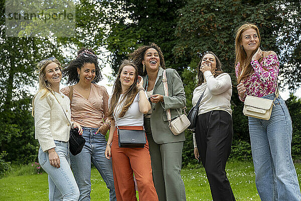 A group of six smiling women standing outdoors in a grassy area  each dressed in casual to smart casual attire  enjoying a moment together under a cloudy sky.