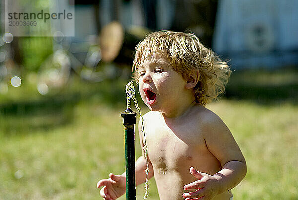 Shirtless toddler joyfully plays with water from a sprinkler on a sunny day.