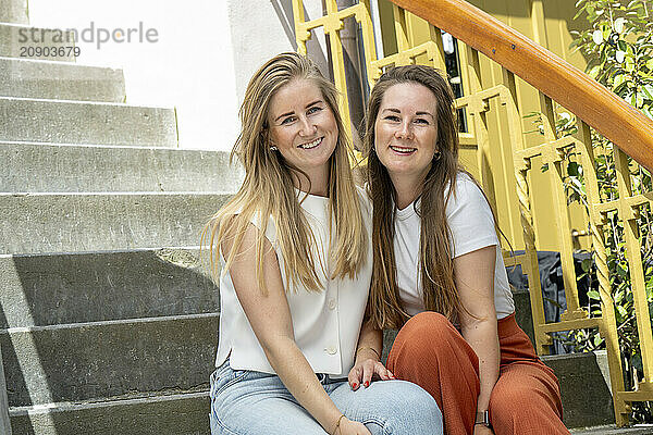 Two smiling women sitting together on steps with a yellow handrail in the background.