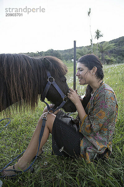 A woman in a floral dress sits on grass and affectionately touches a brown horse's muzzle  New Zealand