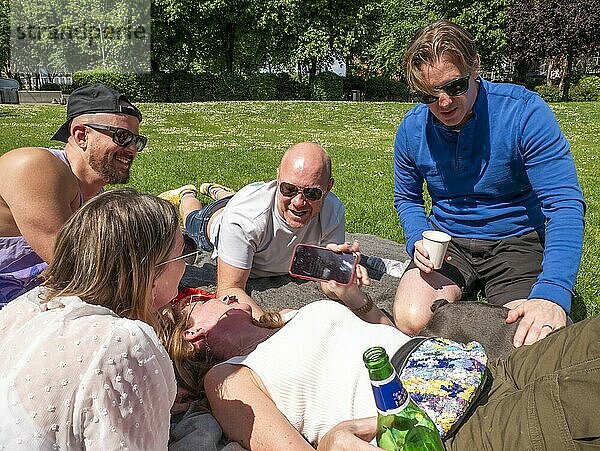 Friends laughing together while lying on a grassy park  with one showing something on a smartphone.