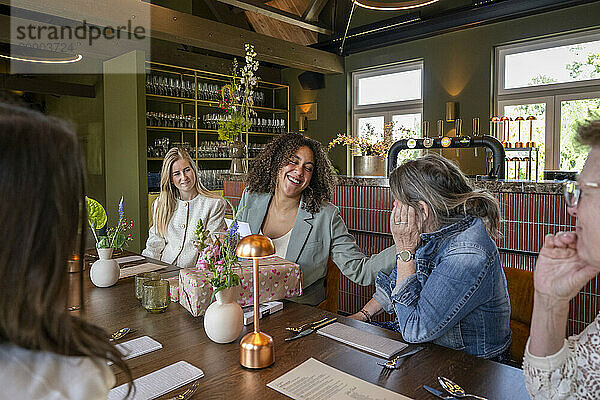 Friends enjoying a surprise gift at a cozy restaurant table  with one woman laughing joyfully as she covers her face.