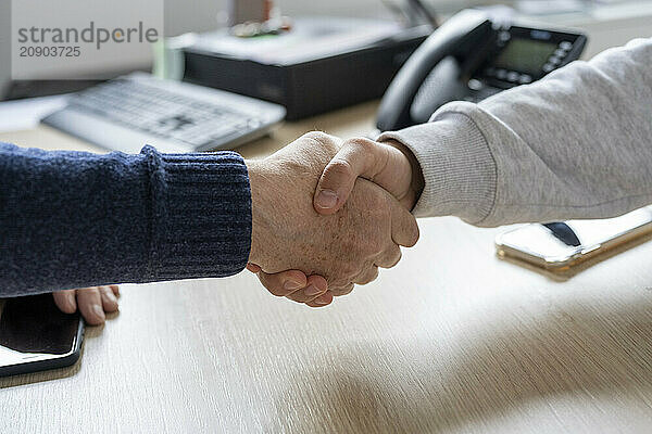 Two individuals engage in a firm handshake over a wooden desk in an office setting