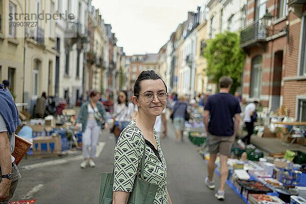 Smiling woman holding a bag stands at a bustling street market with shoppers and stalls in the background.