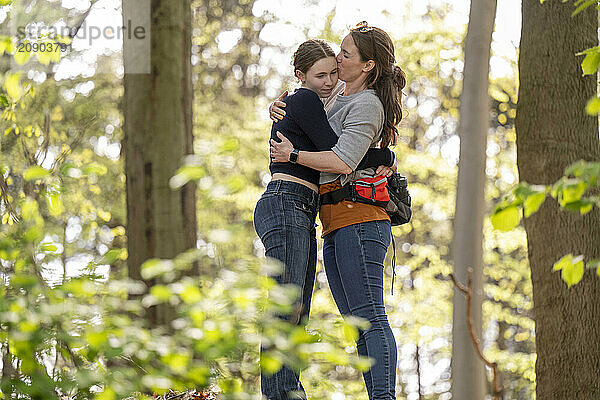 Mother and daughter sharing a tender embrace in a sunlit forest  with the mother affectionately kissing her daughter's forehead.