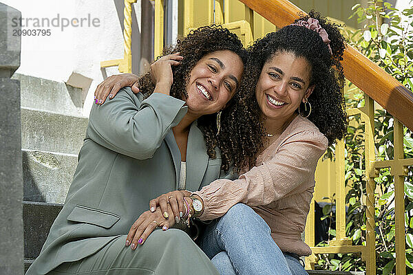 Two smiling women with curly hair embrace warmly while sitting on outdoor steps.