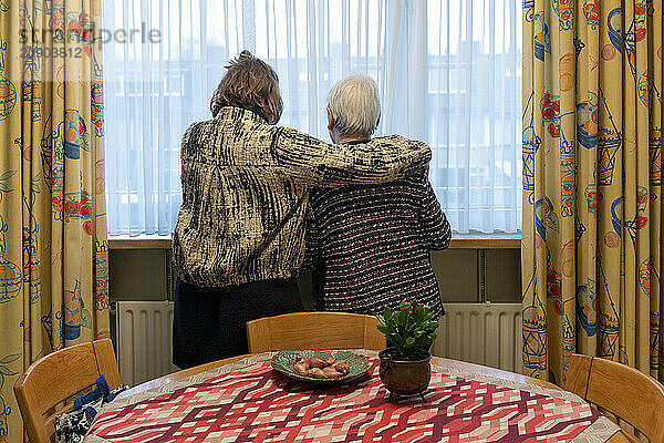 Two elderly women embrace while looking out the window  a cozy room with a flowered curtain and a fruit bowl on the table suggests a homely atmosphere.