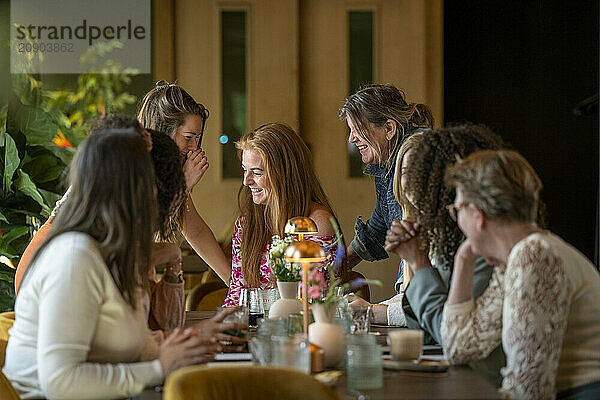 A group of joyful women sharing a laugh around a table beautifully set with flowers and glasses  in a room with natural light.