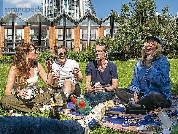 Group of friends enjoying a picnic on a sunny day with drinks and snacks  sitting on a blanket in a grassy park  laughing and conversing with contemporary buildings in the background.