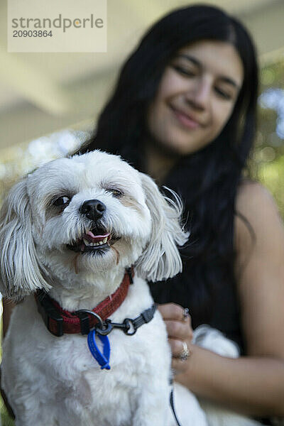 A smiling young woman cuddling her happy white dog in a sunny garden setting.