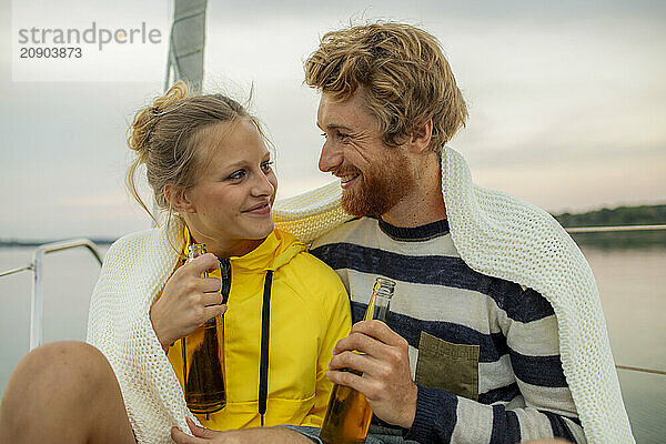 Smiling couple sharing an intimate moment wrapped in a blanket with a bottle  on a boat at sunset