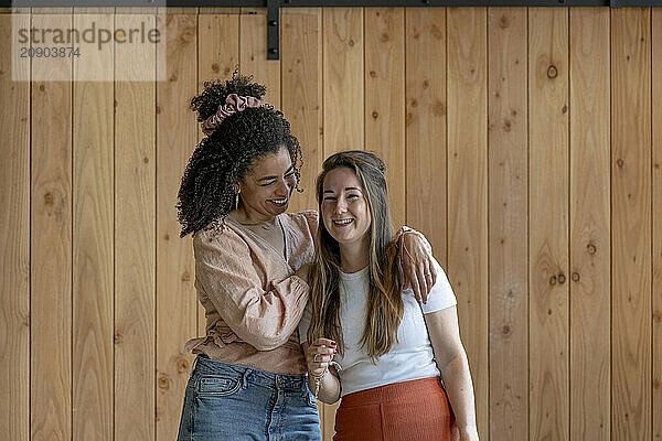 Two women are standing together  one with her arm around the other  both smiling and posing in front of a wooden wall.