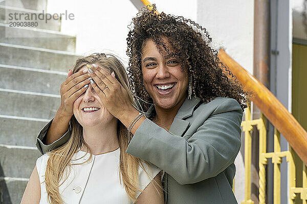 Smiling woman with curly hair covering another woman's eyes for a surprise on steps outdoors.