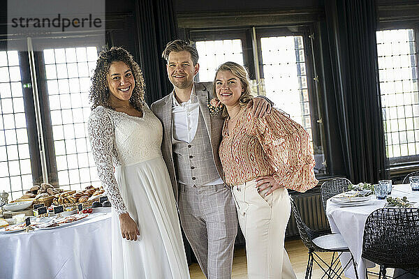 Smiling bridal couple with a guest posing in a banquet hall with a table filled with food.