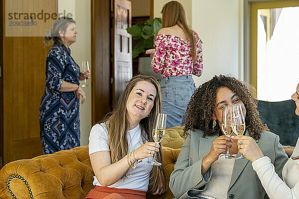 Two women smiling and toasting with glasses of white wine while sitting on a couch  with two other women standing in the background in a cozy home setting.