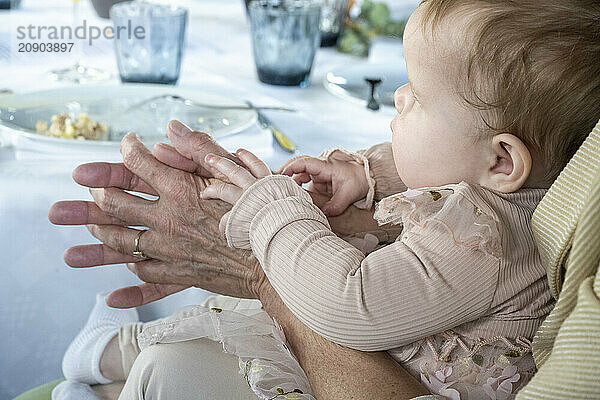 An infant in a pink outfit is held in the arms of a senior  whose wrinkled hands gently grasp the baby's tiny ones at a dining table.