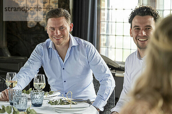 Two smiling men enjoying a meal with wine glasses at an elegantly set table  with a focus on the man in the foreground.