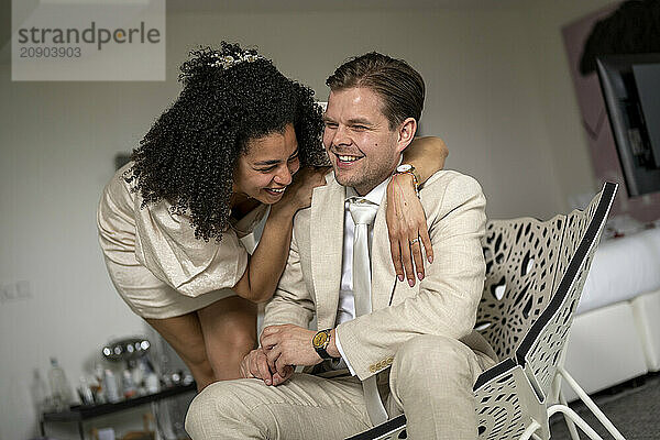 Happy couple laughing and embracing while sitting on a chair in a well-lit room.