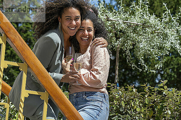 Two women embracing and laughing on a sunny day outdoors with a glass of wine.