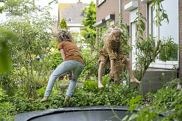 Two children jump joyfully on a trampoline in a backyard garden surrounded by greenery