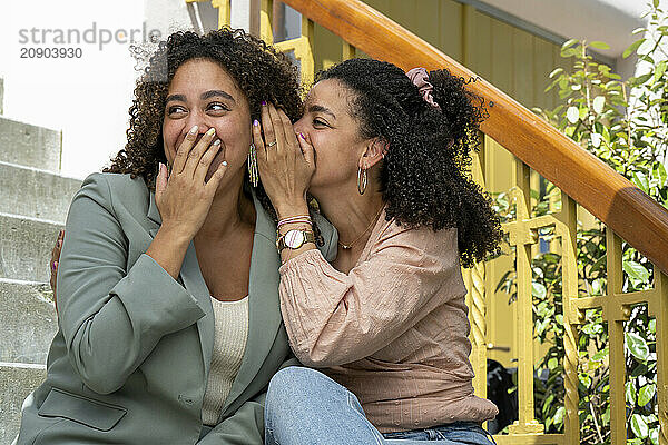 Two women share a moment of laughter and whispering on outdoor steps.