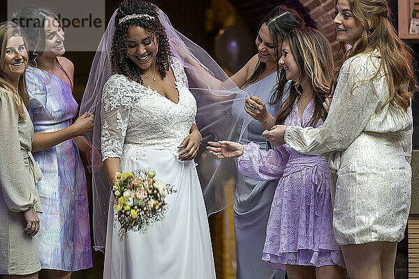 A radiant bride in a white lace dress smiles joyfully  surrounded by her bridesmaids in pastel attire  as they admire her gown and share a special moment together.