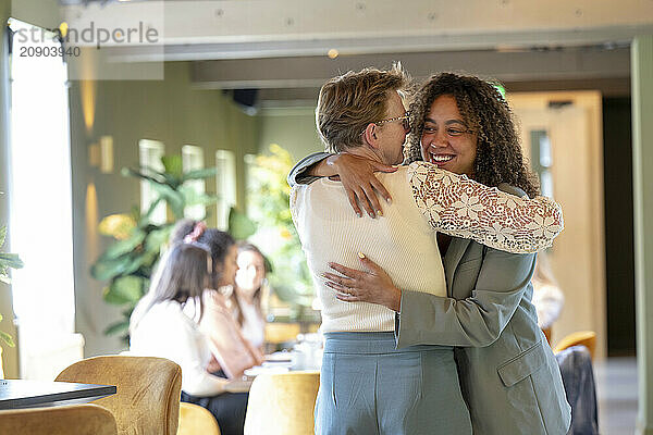 Two women embracing warmly in a sunny caf? setting with other guests in the background.