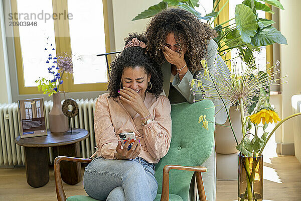 Two women share a joyful moment while looking at a smartphone together in a cozy  sunlit room with plants and stylish decor.