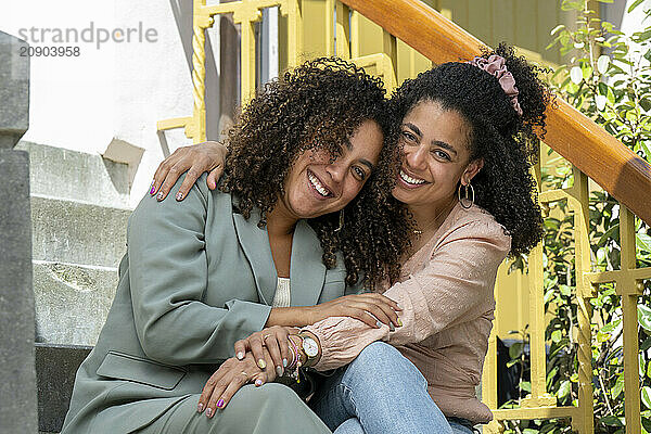 Two smiling women embracing on outdoor steps  both with curly hair and stylish attire.