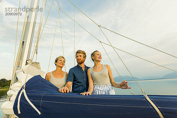 Three friends enjoy a sunny day on a sailboat with a clear blue sky and tranquil sea in the background.