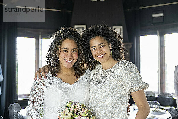 Two smiling women embracing and posing for a photo in an interior setting  one holding a bouquet of flowers.