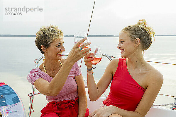 Two women enjoying drinks and smiling at each other on a boat with a calm lake in the background.