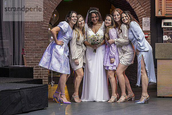 A joyful bride in a white dress and her bridesmaids in pastel outfits pose for a photo at an indoor wedding venue.