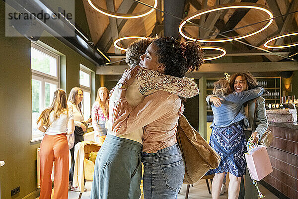 Two women embracing in a joyful hug with others socializing in the background in a warmly lit room.