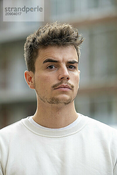 A portrait of a young man with tousled hair and slight stubble wearing a white t-shirt  standing in front of a blurred building background.