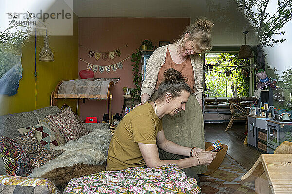 A smiling woman showing another woman something on a smartphone in a cozy living room with plants and bohemian decor.