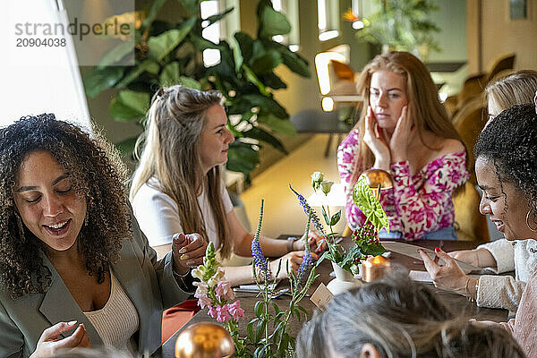 A group of women enjoying a conversation at a brightly lit restaurant table decorated with fresh flowers.