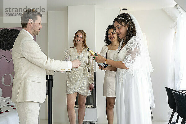 A man in a suit pours champagne for a smiling bride and two women celebrating indoors.