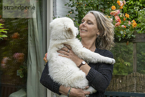 A smiling woman holding a fluffy white dog in her arms with colorful flowers in the background.