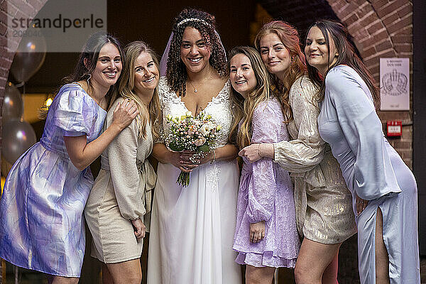 A smiling bride in a white dress holding a bouquet stands surrounded by five joyful bridesmaids in pastel dresses at an indoor celebration.