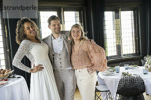 Three friends dressed in formal wear smile together at an elegant indoor event.