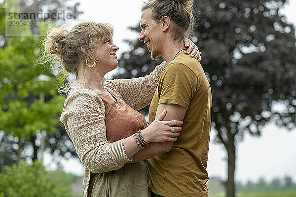 Two women embracing and smiling at each other in a park with trees in the background.